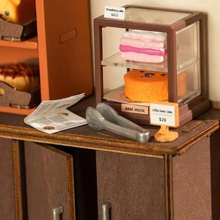 Two miniature cakes sit behind clear panels beside tiny tongs.